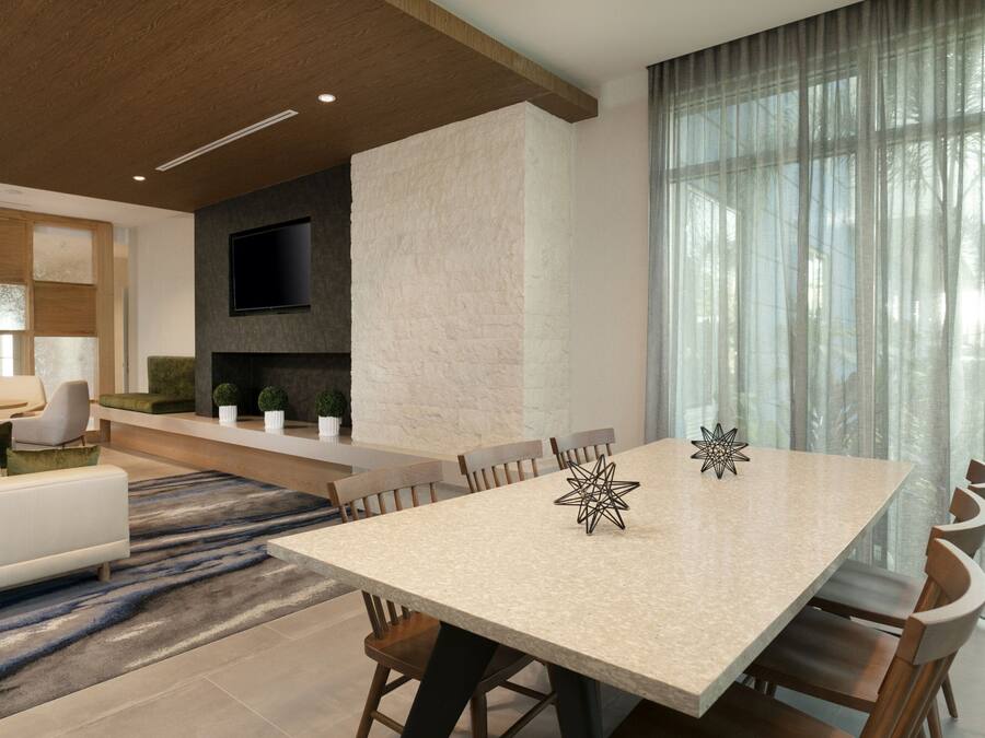 A modern living room with a large dining table, comfortable seating, a flat-screen TV mounted on a textured wall, and a large window with sheer curtains.