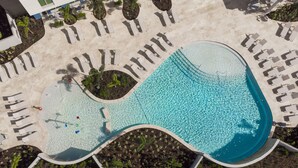 Vue aérienne d’une piscine au design original, avec des formes ondulées, entourée de chaises longues et de petits palmiers.