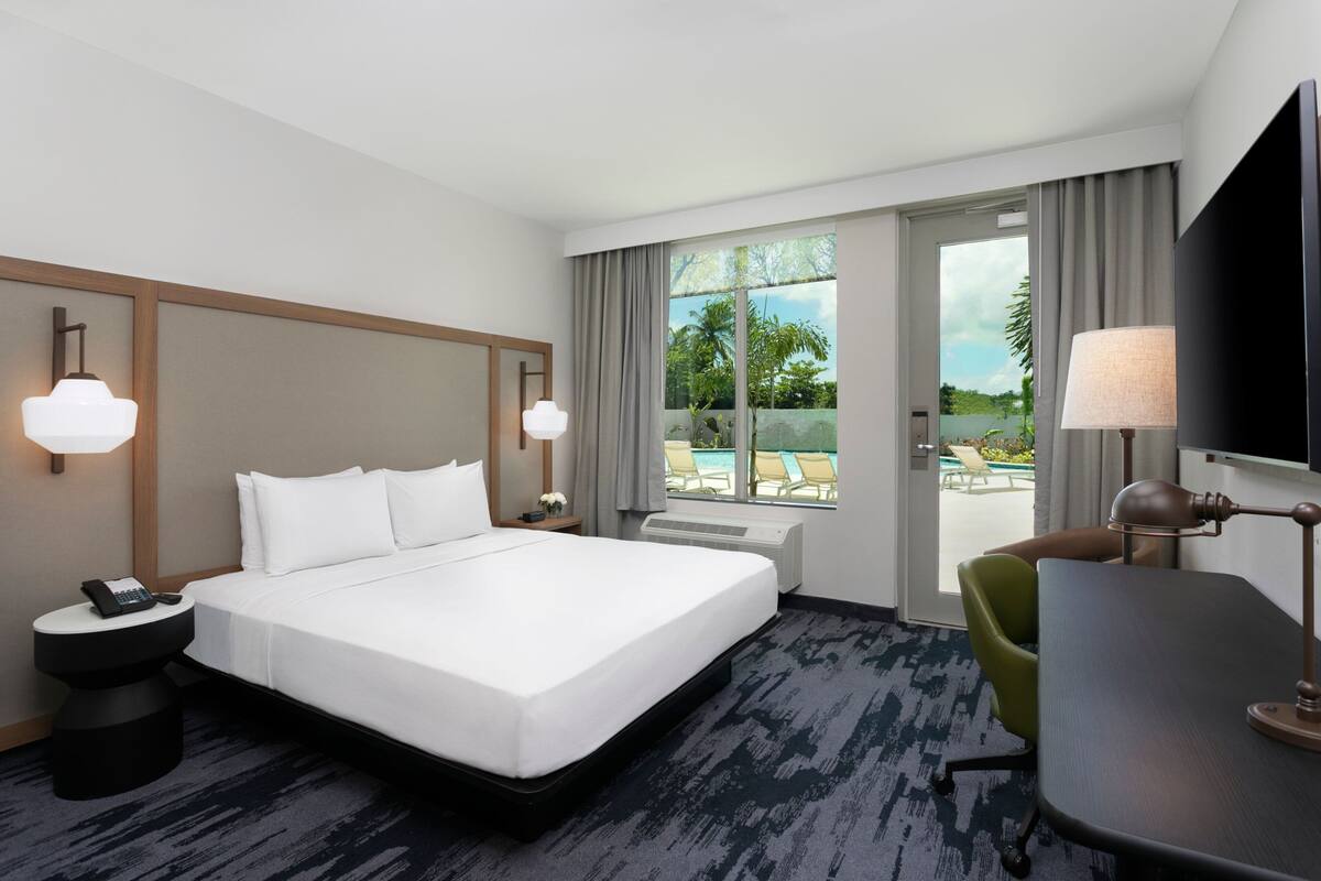 A hotel room with a large bed, a desk, a chair, a TV, and a view of a pool and greenery.