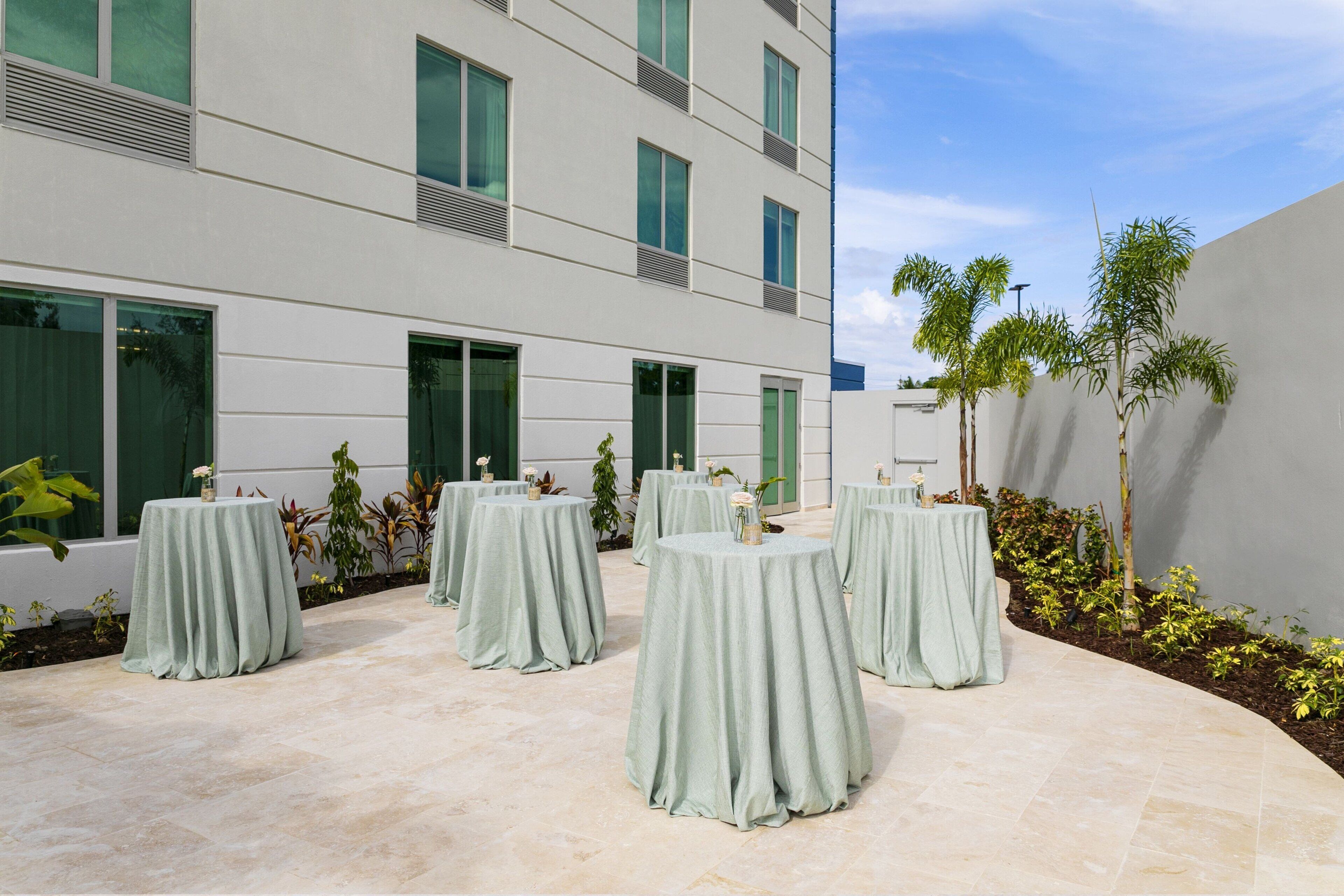 Un patio con mesas redondas cubiertas con manteles verde claro, rodeado de plantas en macetas y un edificio moderno con amplios ventanales.