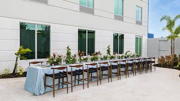 A long table set outdoors with chairs, positioned in front of a modern building with large windows and a tiled floor.
