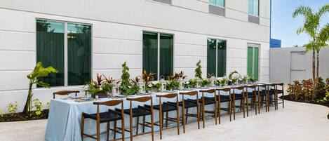 A long table set outdoors with chairs, positioned in front of a modern building with large windows and a tiled floor.