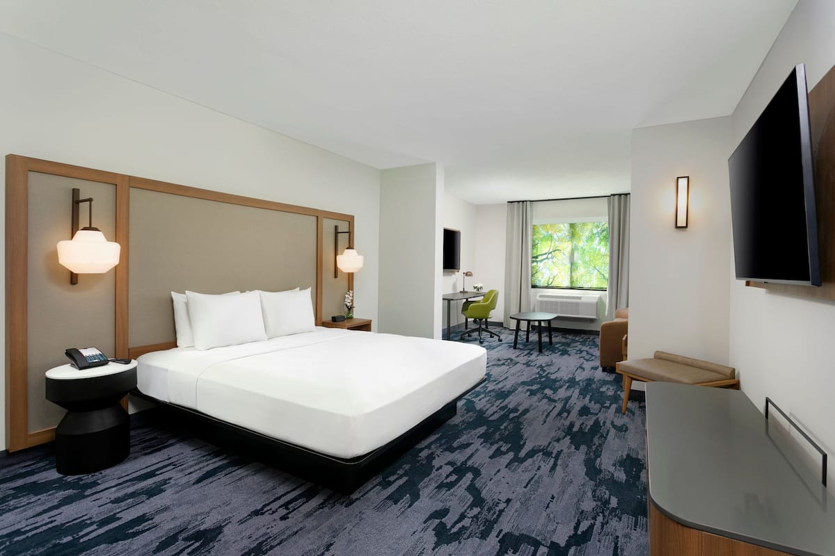 A hotel room with a large bed, bedside tables, a desk, a chair, a TV, and a window with a view of greenery.