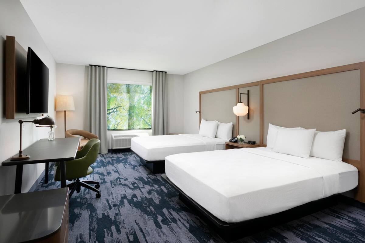 A hotel room with two beds, a desk with a chair, a TV, and a window with curtains.