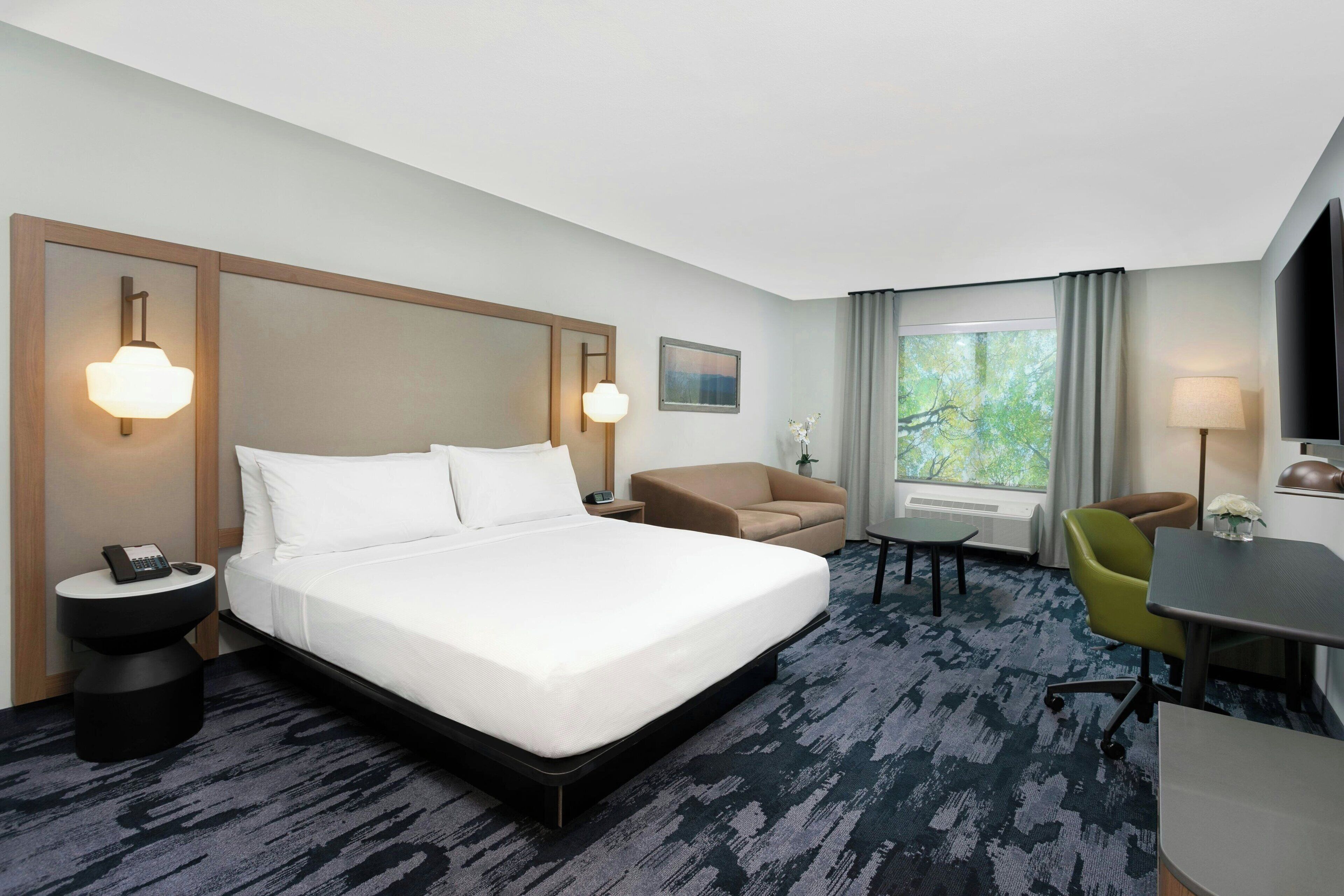 Quarto de hotel com uma cama grande, uma mesa, uma cadeira e um sofá. Há uma janela com cortinas e vista para árvores.
