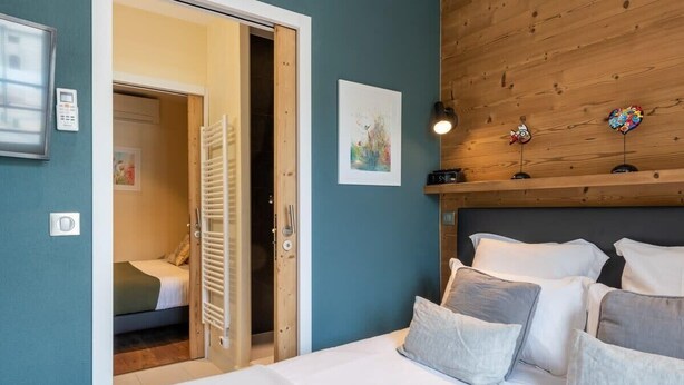 Un dormitorio con cama, cabecero de madera, una puerta que da a otra habitación y una lámpara fijada a la pared.
