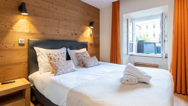 A hotel room with a wooden headboard, a large bed with white bedding, two wall-mounted lamps, and a window with orange curtains. - APPALACHES, Annecy Rent Lodge (Annecy)