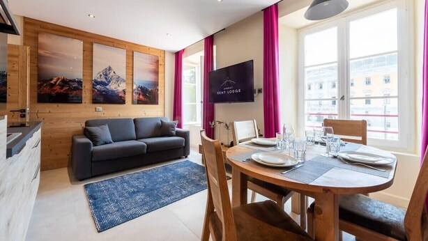 A modern living room with a sofa, dining table, and chairs. There are paintings on the wall, a TV, and a window with curtains. - CAUCASE, Annecy Rent Lodge (Annecy)