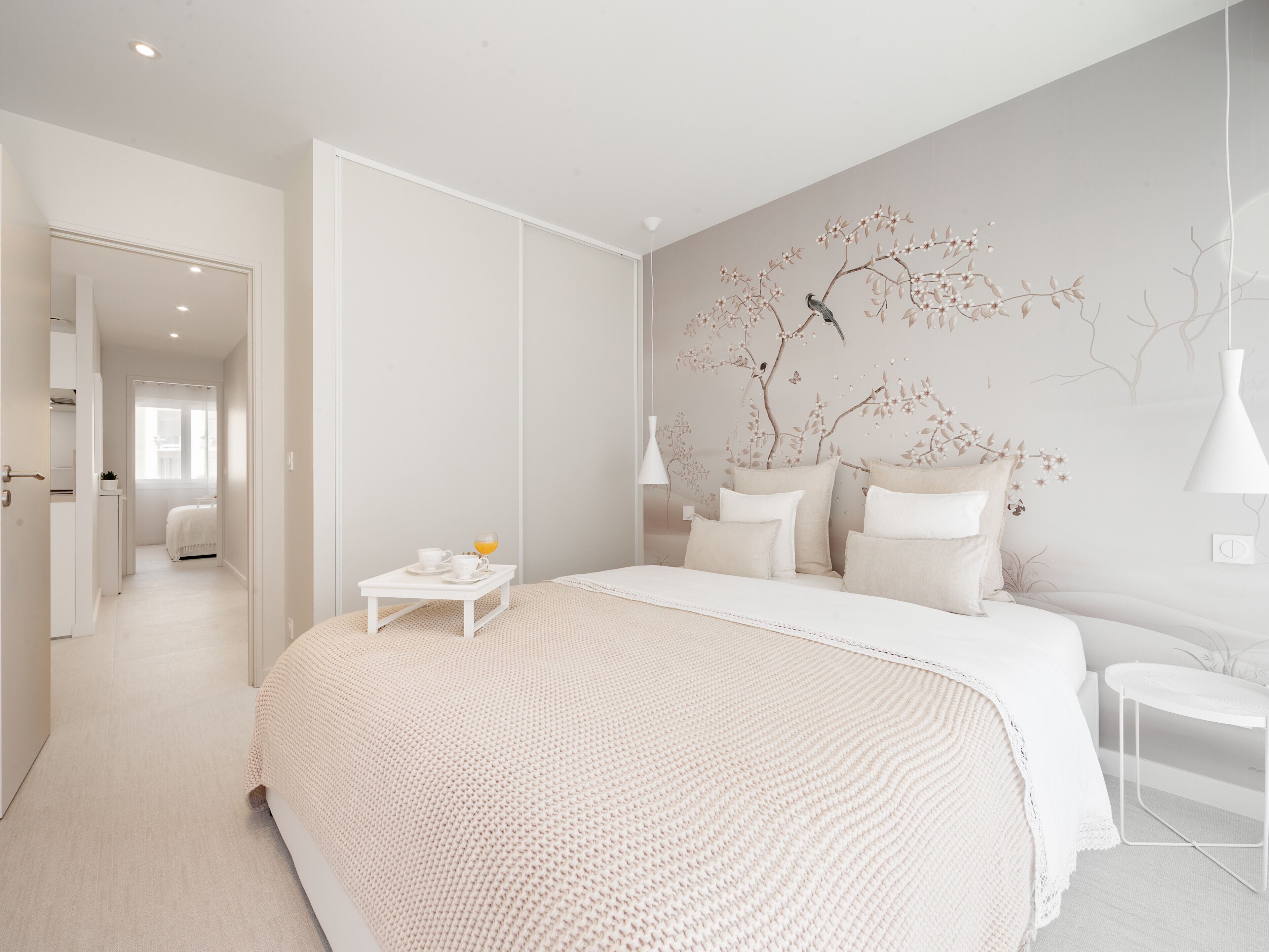 A modern bedroom with a large bed, a small bedside table with a cup and saucer, and a wall mural of a tree.