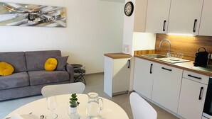 A modern kitchen with white cabinets, a wooden countertop, and a sink. There is a dining table set with plates, glasses, and cutlery. A gray sofa with yellow cushions is in the living area, and a clock is on the wall. - Le Quartz Tresum, in a modern residence near the lake, garage included (Annecy)