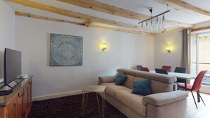 Een woonkamer met een bank, een televisie, een houten kast, een kroonluchter, en een eethoek met stoelen en een tafel.