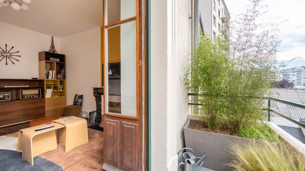 A cozy living room with a wooden bookshelf, a small table, and a door leading to a balcony with a view of buildings and greenery. - Le Cosy d'Aléry - Charming 3 room apartment near downtown (Annecy)