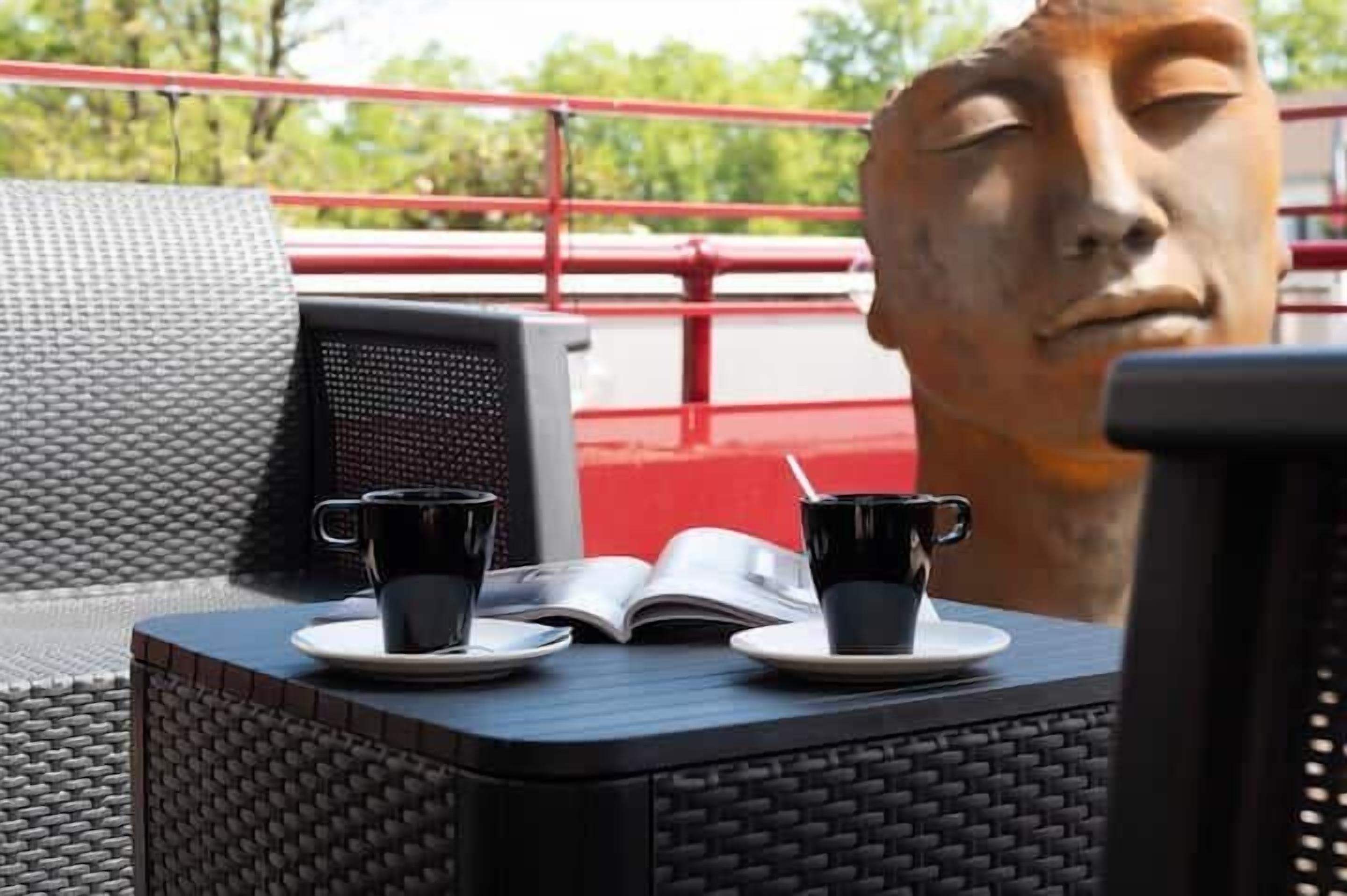 A table with two cups and an open book, a wicker chair, and a red railing in the background.