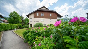 Un bâtiment à deux étages, avec un toit brun et des murs de couleur beige, entouré d’une végétation luxuriante et de fleurs roses.
