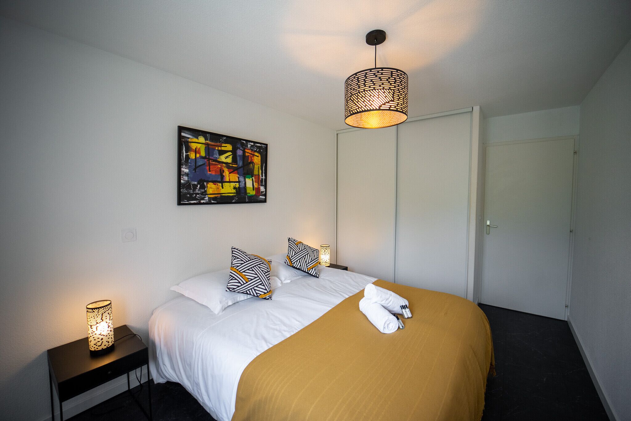 Una cama bien hecha con ropa blanca y un plaid marrón, una lámpara colgante moderna y un cuadro abstracto de colores en la pared.