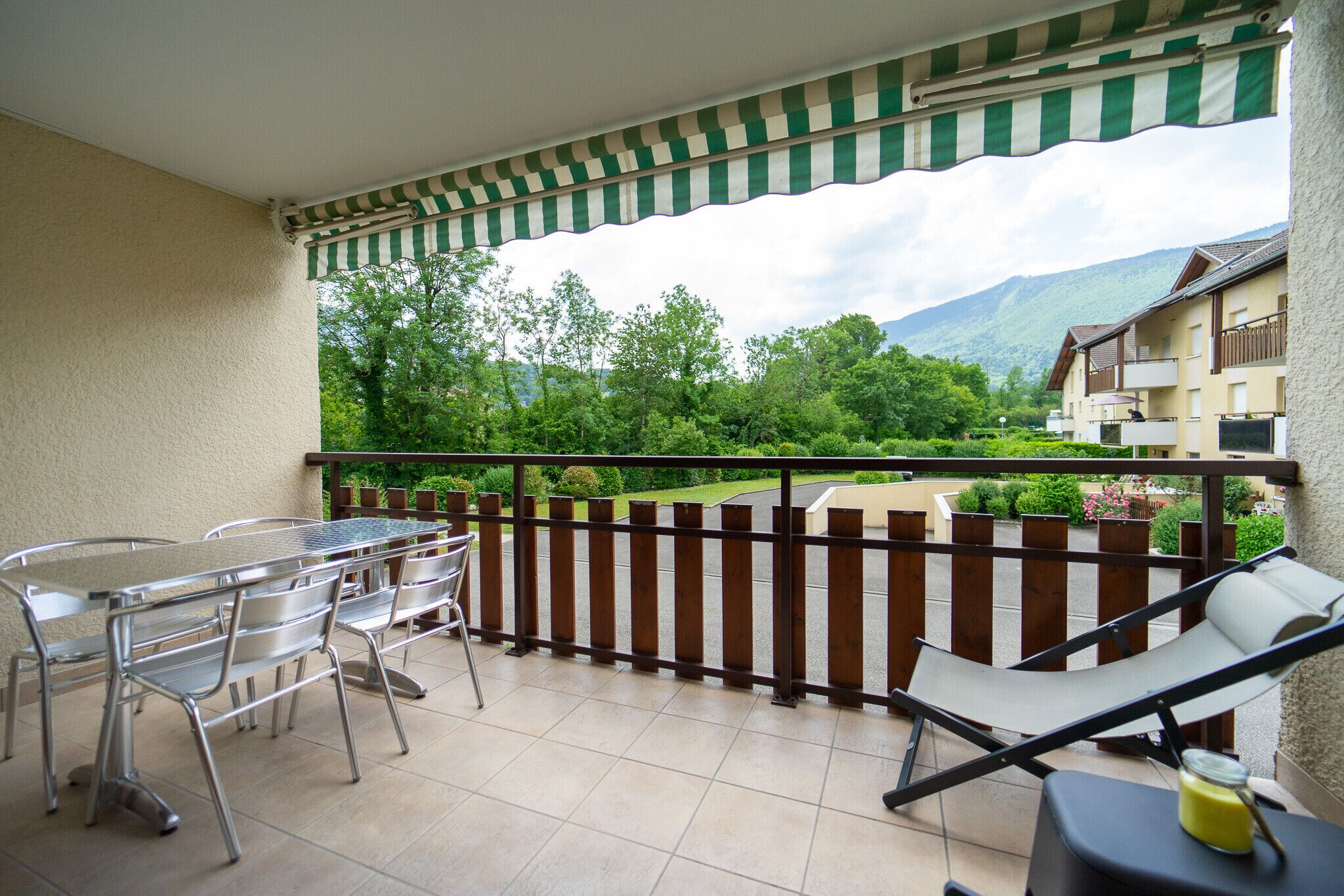 Ein Balkon mit einem Metalltisch und Stühlen, einem gepolsterten Stuhl und Blick auf Grünflächen und Gebäude im Hintergrund.