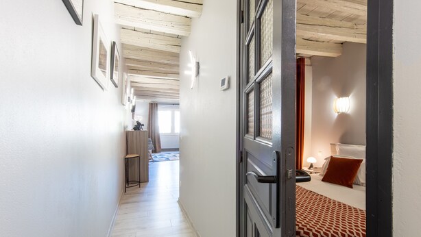 Un pasillo que conduce a un dormitorio con cama, mesita de noche y lámpara. Se aprecia una ventana con cortinas, una puerta y una lámpara fijada a la pared.
