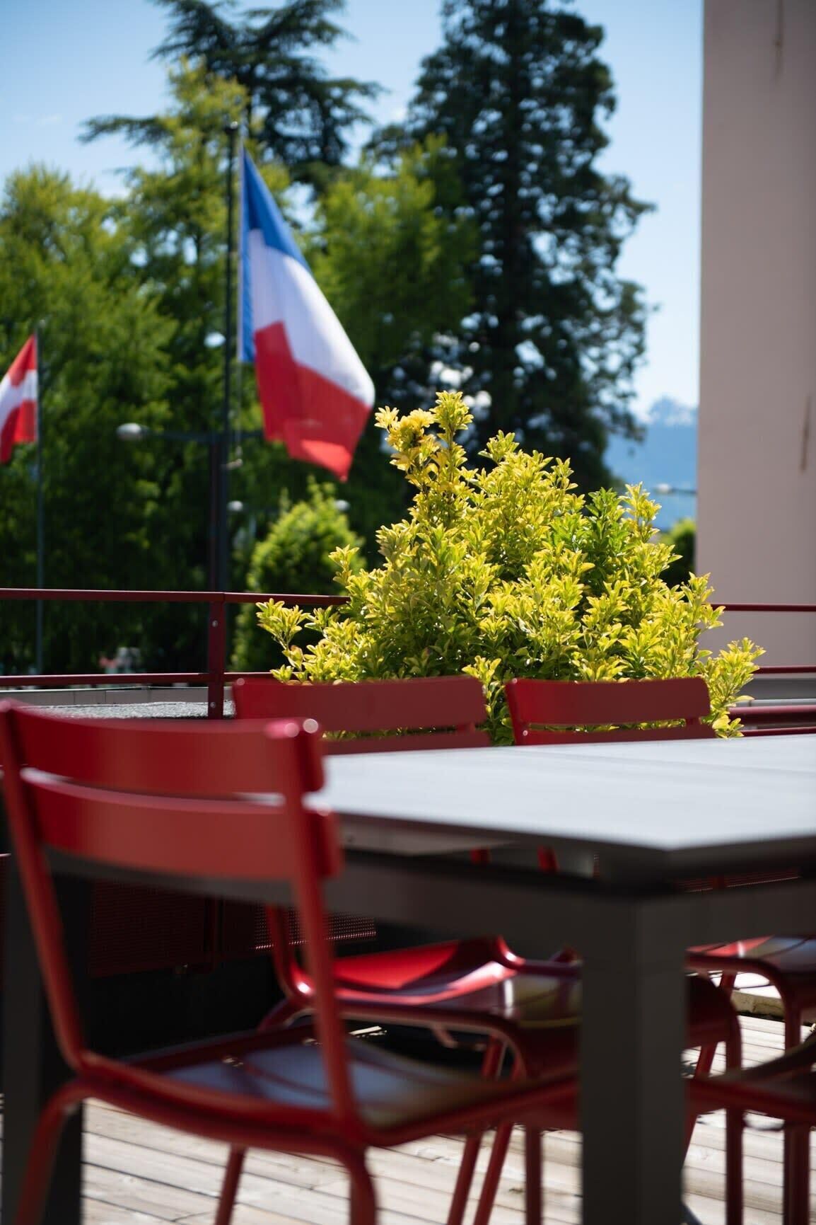 A patio area with red chairs and a table, a small bush, and flags in the background.