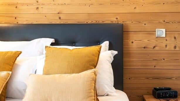 A neatly made bed with white and mustard pillows, a dark headboard, and a wooden wall with a wall-mounted lamp and a small table with a clock.