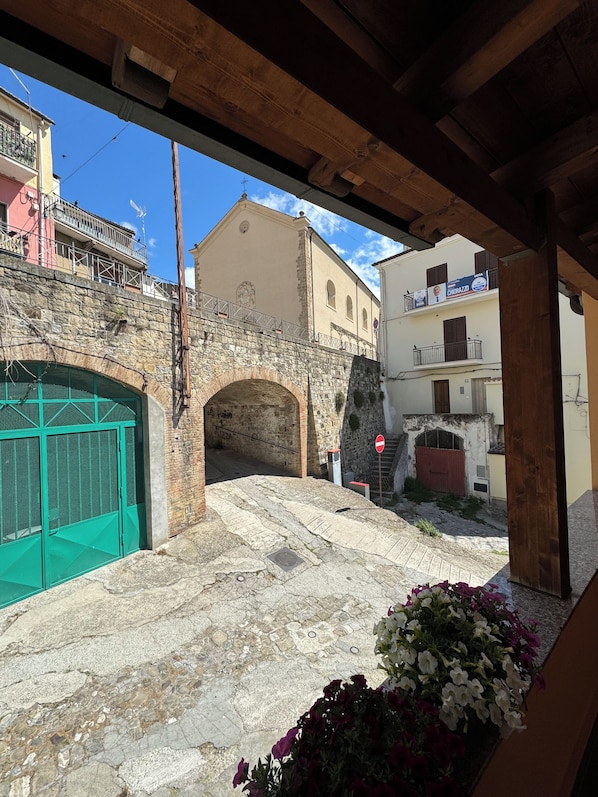 A stone archway leading to a courtyard with a green gate, a flower bed, and buildings with balconies and signage.