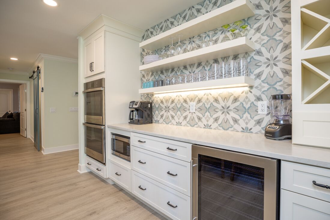 A modern kitchen with white cabinetry, built-in ovens, a microwave, and a dishwasher. There are shelves above the countertop holding glassware and a blender.