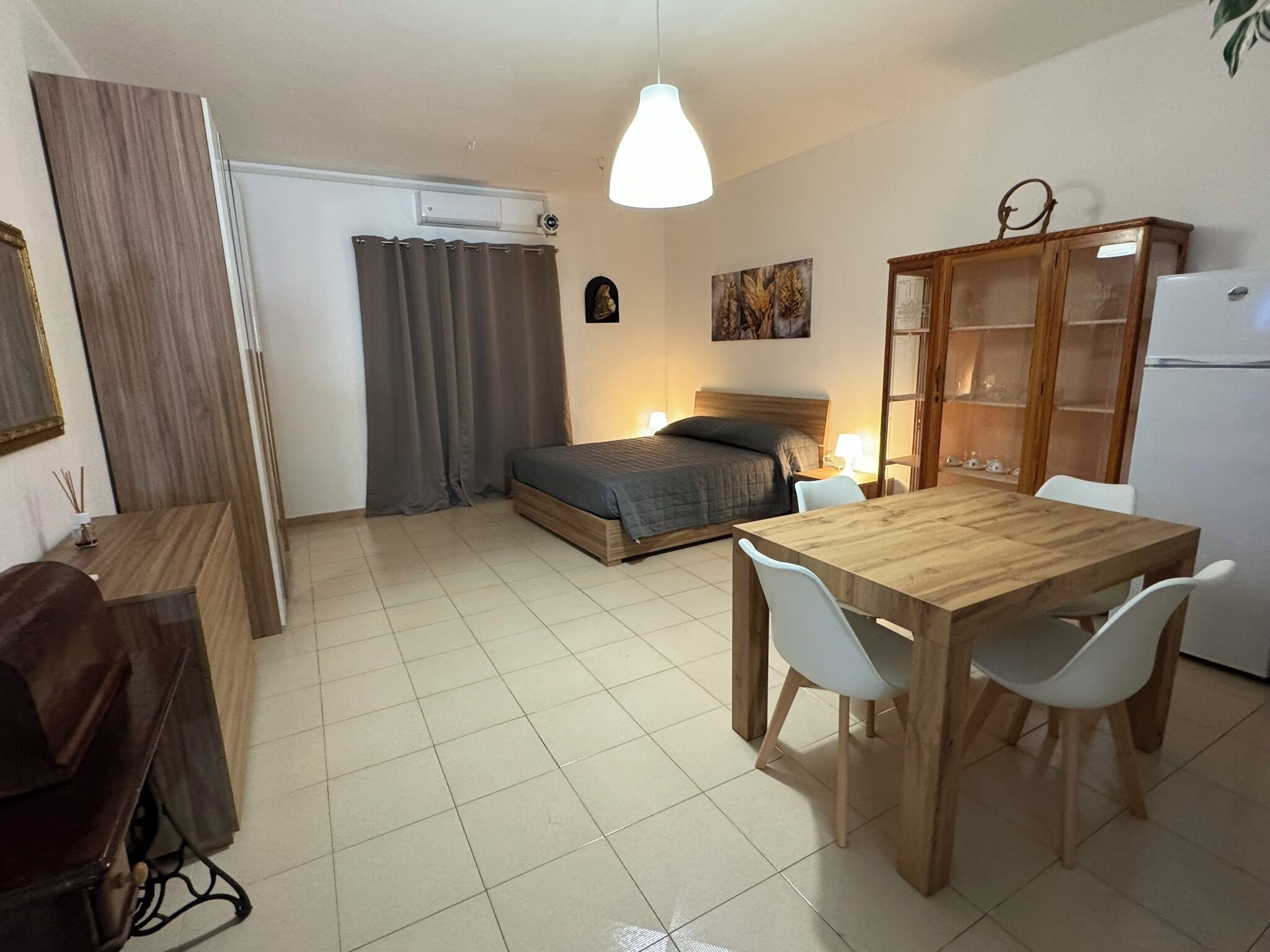 A compact room with a bed, a wooden dining table with chairs, a small kitchenette, and a cabinet.