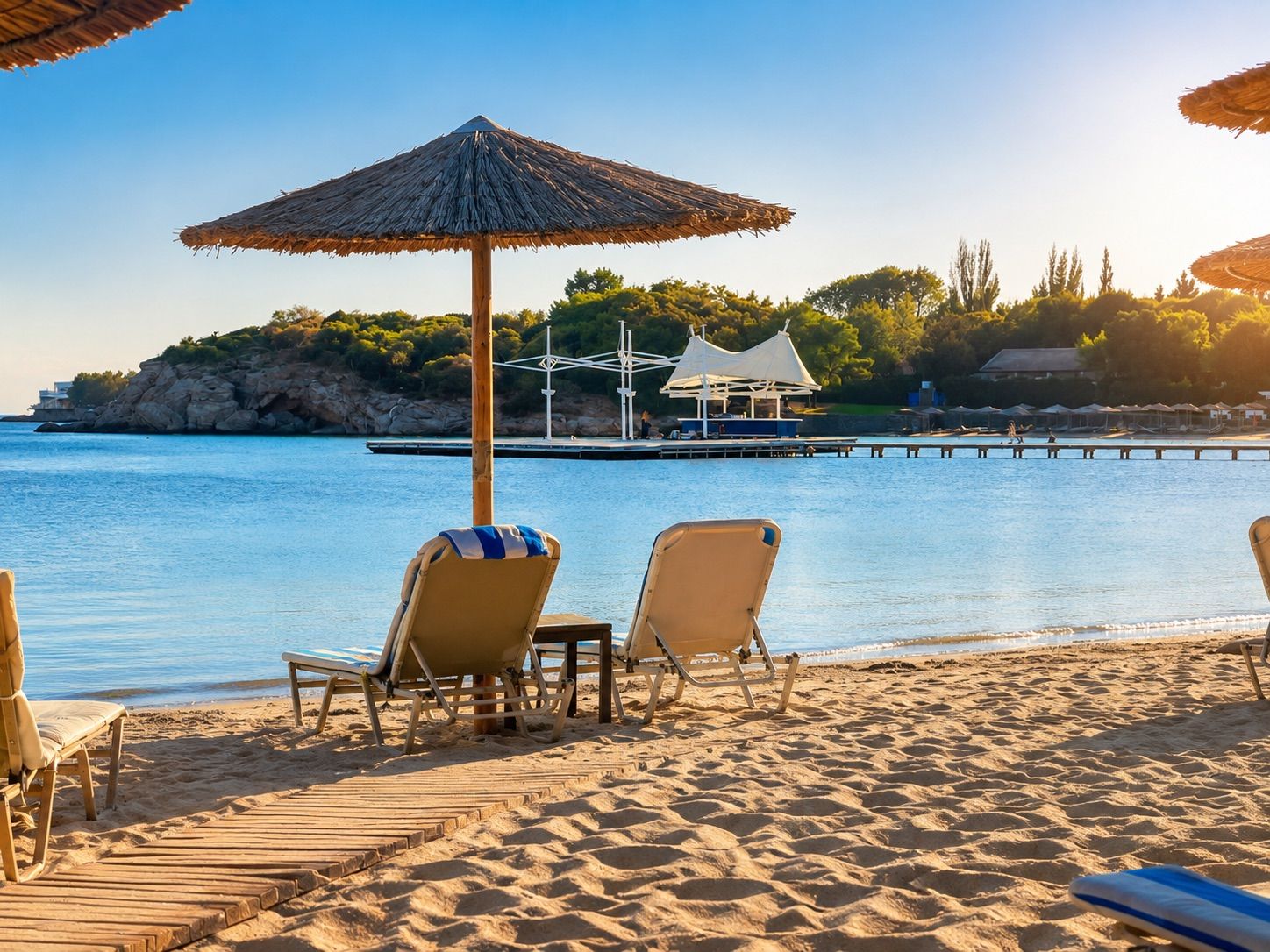 A beach with lounge chairs and a thatched umbrella, a wooden pier, and a rocky coastline.