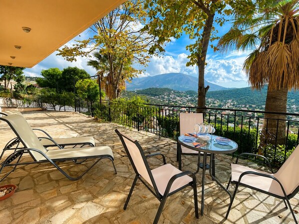 An outdoor terrace with stone flooring, a table with chairs, and a view of a mountainous landscape.