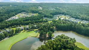 An aerial view of a golf course with a lake, surrounded by dense forest and residential buildings.