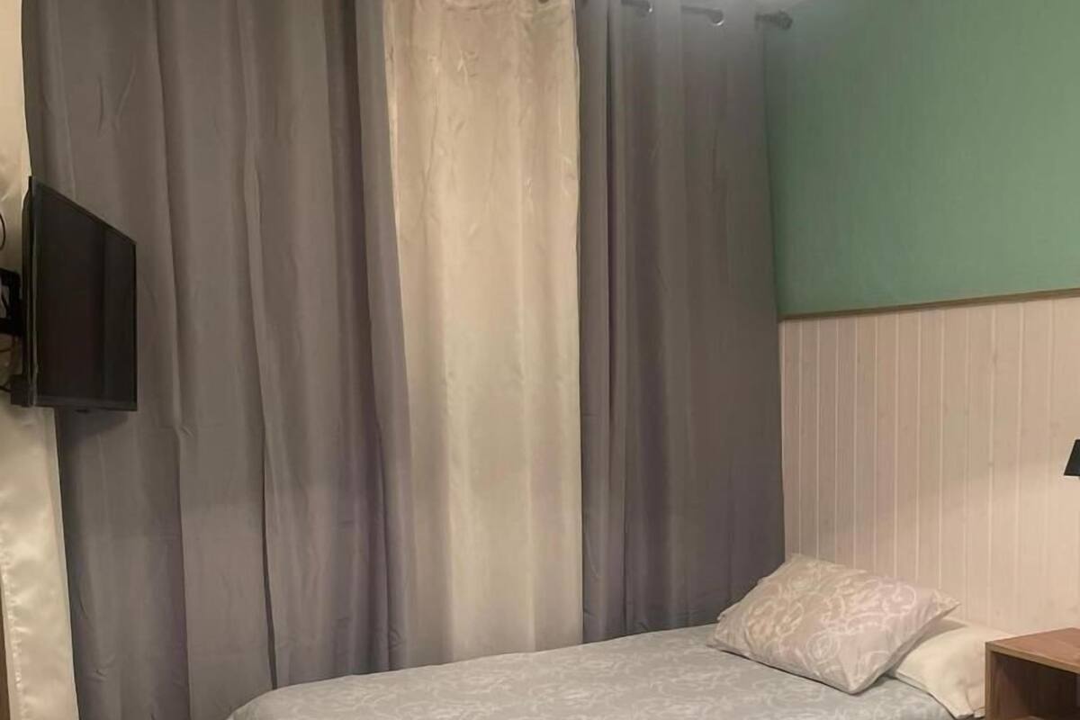 Een slaapkamer met een bed, gordijnen, een aan de muur bevestigde televisie en een nachtkastje.
