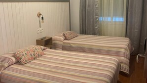 Una habitación con dos camas, una mesita de noche, una lámpara y un ventanal con cortinas.