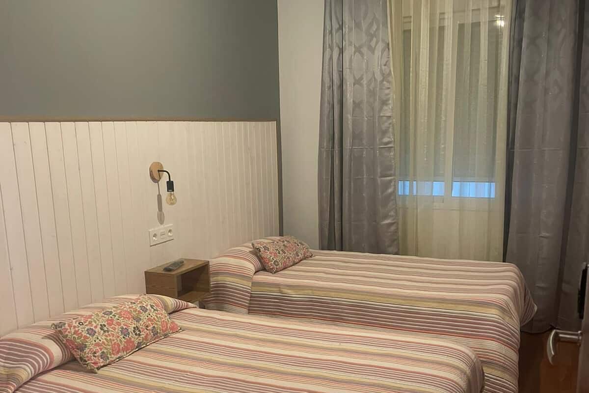 Een kamer met twee bedden, een nachtkastje, een lamp en een raam met gordijnen.