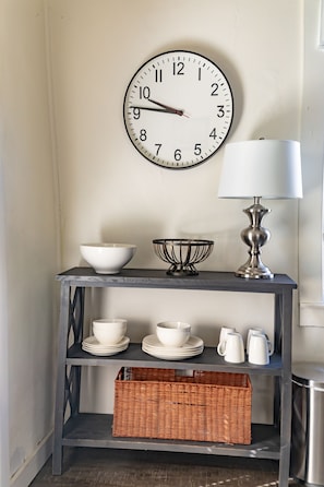 A wall clock, a table lamp, and a shelf with bowls, plates, and a basket. - The Jesse: Cottage in Historic Downtown Pensacola (Pensacola)