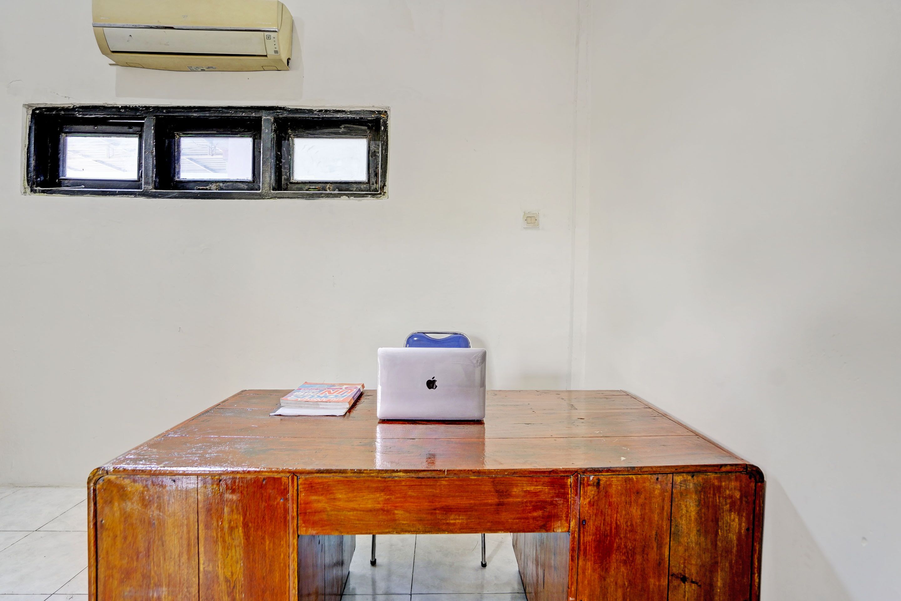 Una mesa de madera con una computadora portátil y un libro, una ventana con marcos negros, y un aire acondicionado en la pared.