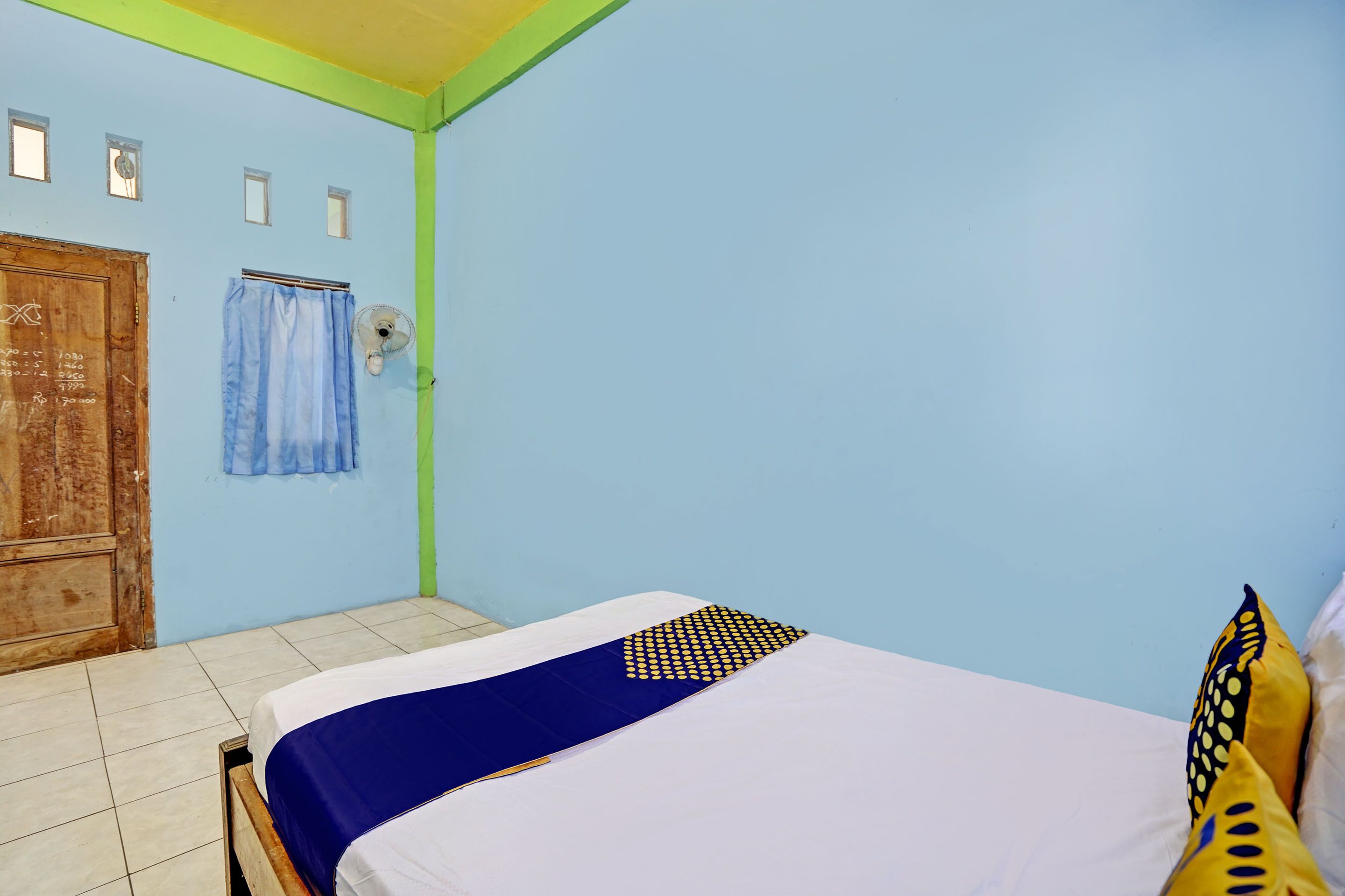 A room with a bed, a wooden door, a window with curtains, and a wall-mounted fan.