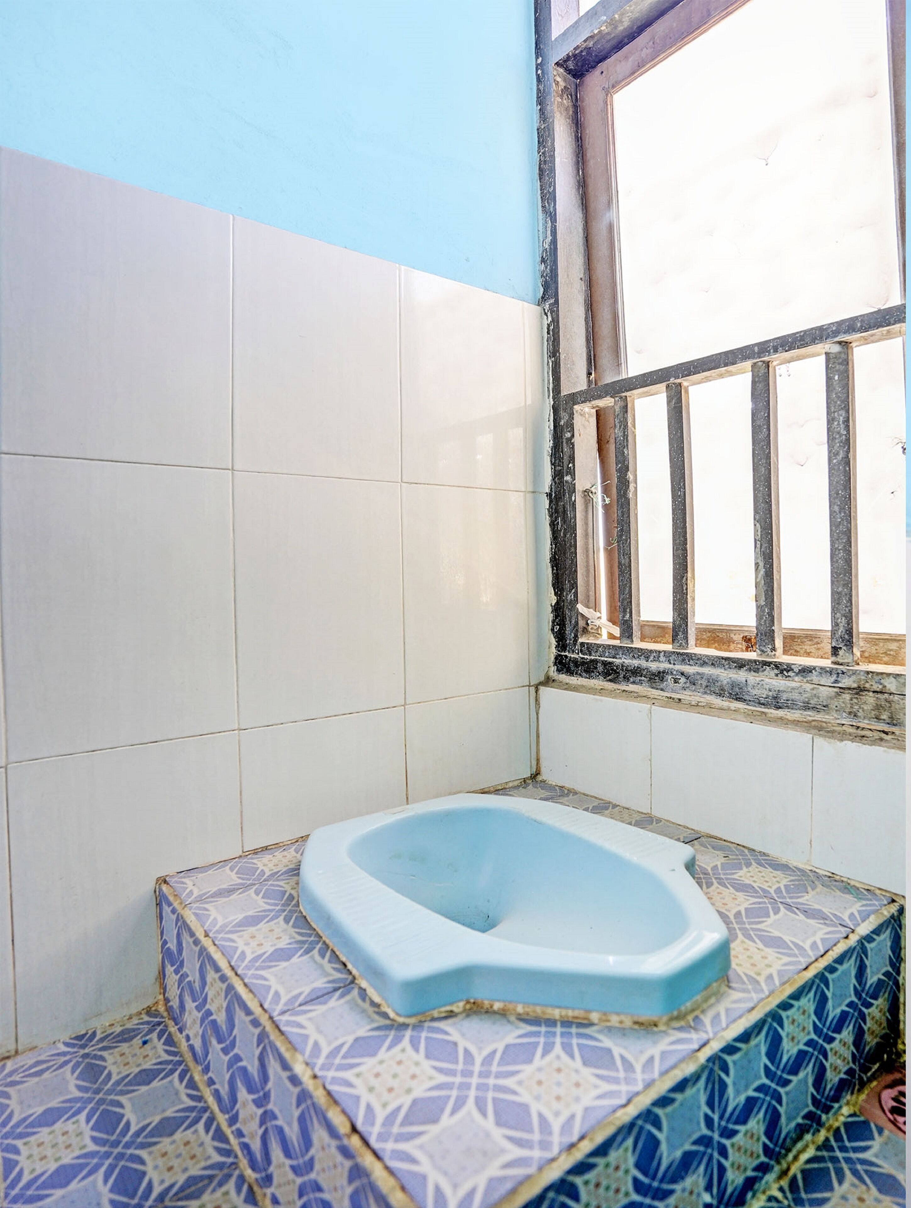 Un baño pequeño con inodoro azul, azulejos blancos y azules con diseño, y una ventana con barrotes.