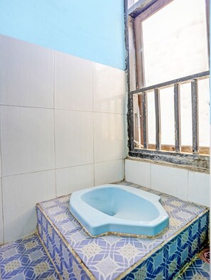 Un baño pequeño con inodoro azul, azulejos blancos y azules con diseño, y una ventana con barrotes.