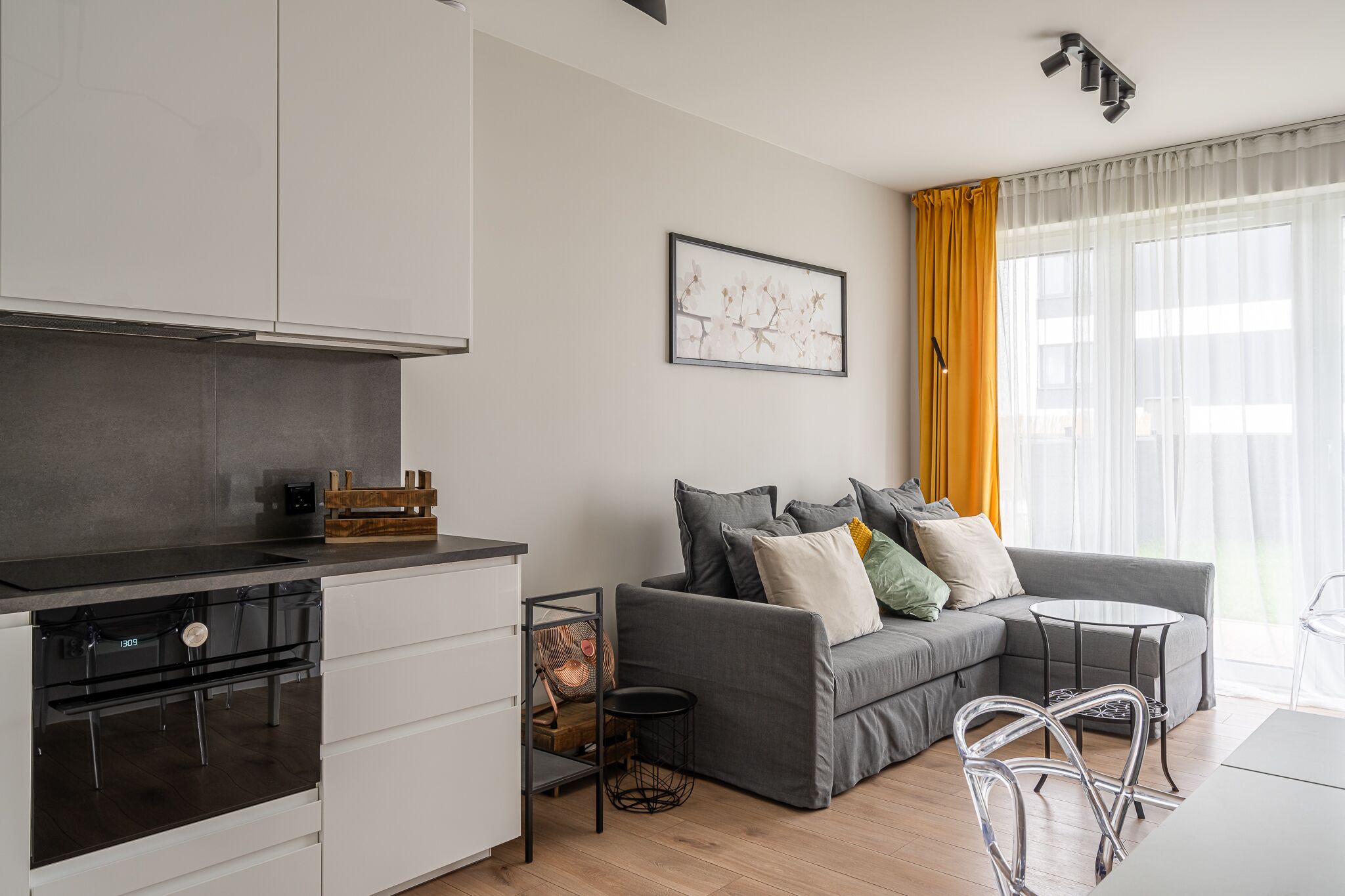 A modern kitchen with white cabinets and a built-in oven, a grey sofa with cushions, a small round table, and a clear chair.