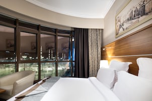 A hotel room with a large bed, a sitting area, and a view of a cityscape through large windows.