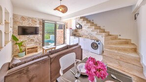 A modern living space with a stone wall, a flat-screen TV, a sofa, a dining table with pink flowers, and a kitchen with a washing machine. - Nouba Villas (Kas)