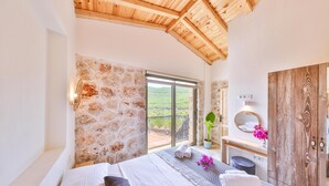 A bedroom with a stone wall, a large window, a wooden ceiling, a bed, a vanity with a mirror, and a potted plant. - Nouba Villas (Kas)