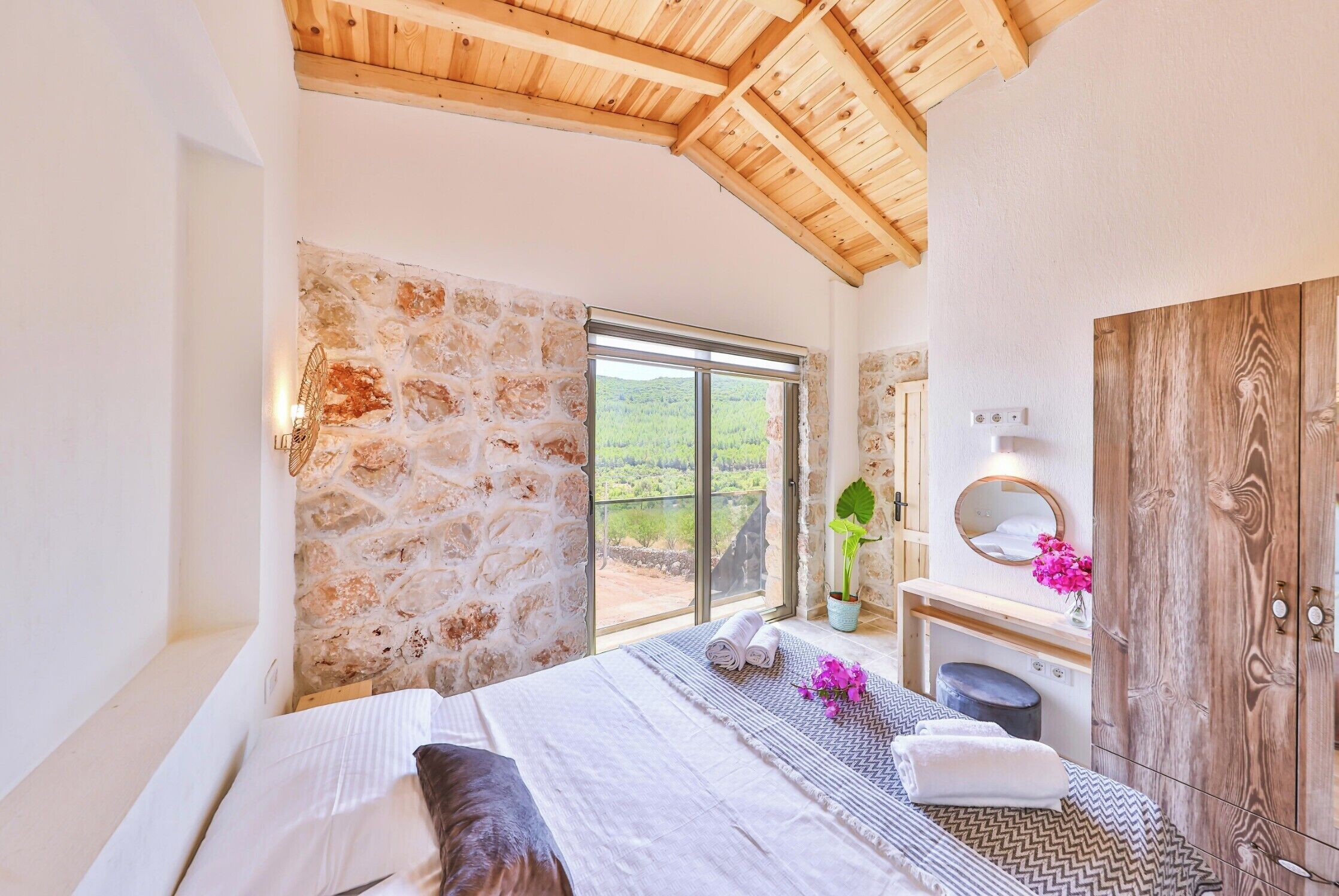 Un dormitorio con pared de piedra, techo de madera, una ventana grande, cama, mesita de noche, espejo y armario.
