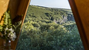 A view through a window of a lush green landscape with a hill and a cliff in the distance. - K4 Kudhva Treehouse Cabin With sea View (Tintagel)