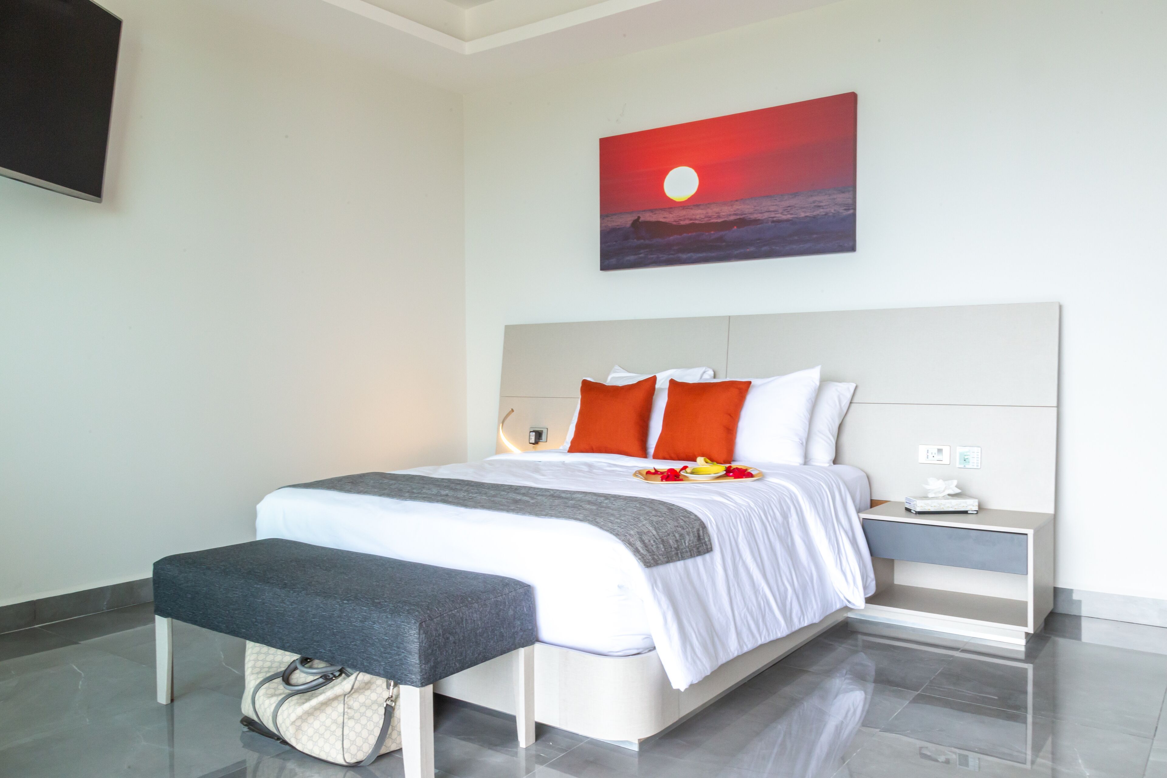 A modern hotel room with a large bed, a bench, a nightstand, and a painting of a sunset.