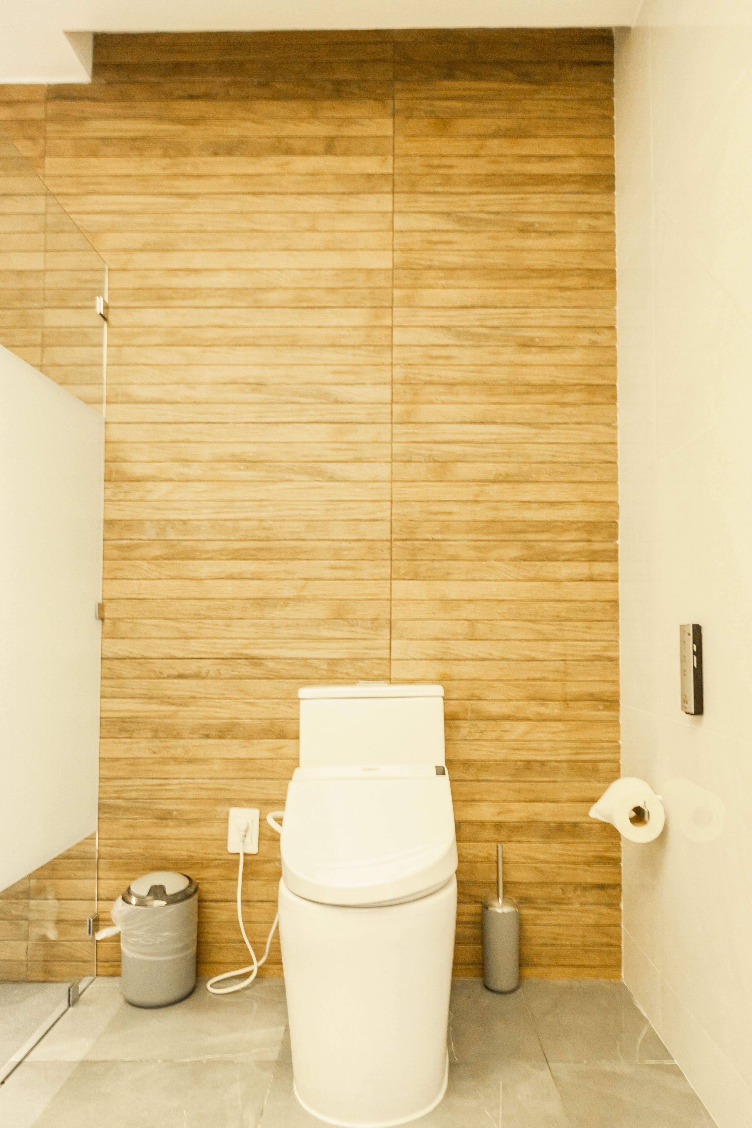 A modern bathroom with a wooden wall, a toilet, a trash bin, and a hand dryer.