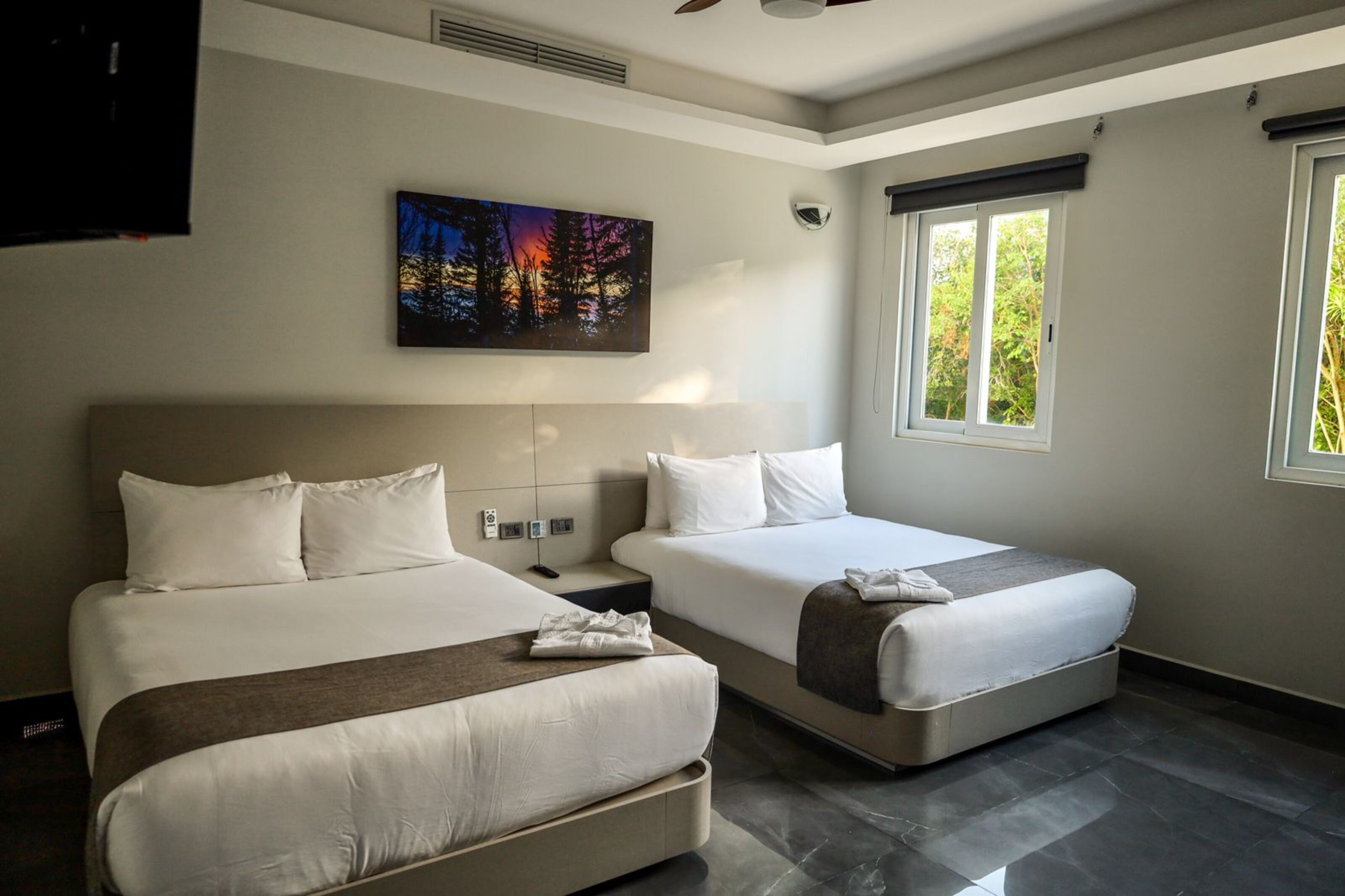A hotel room with two beds, a painting on the wall, a window with a view of trees, and a ceiling fan.
