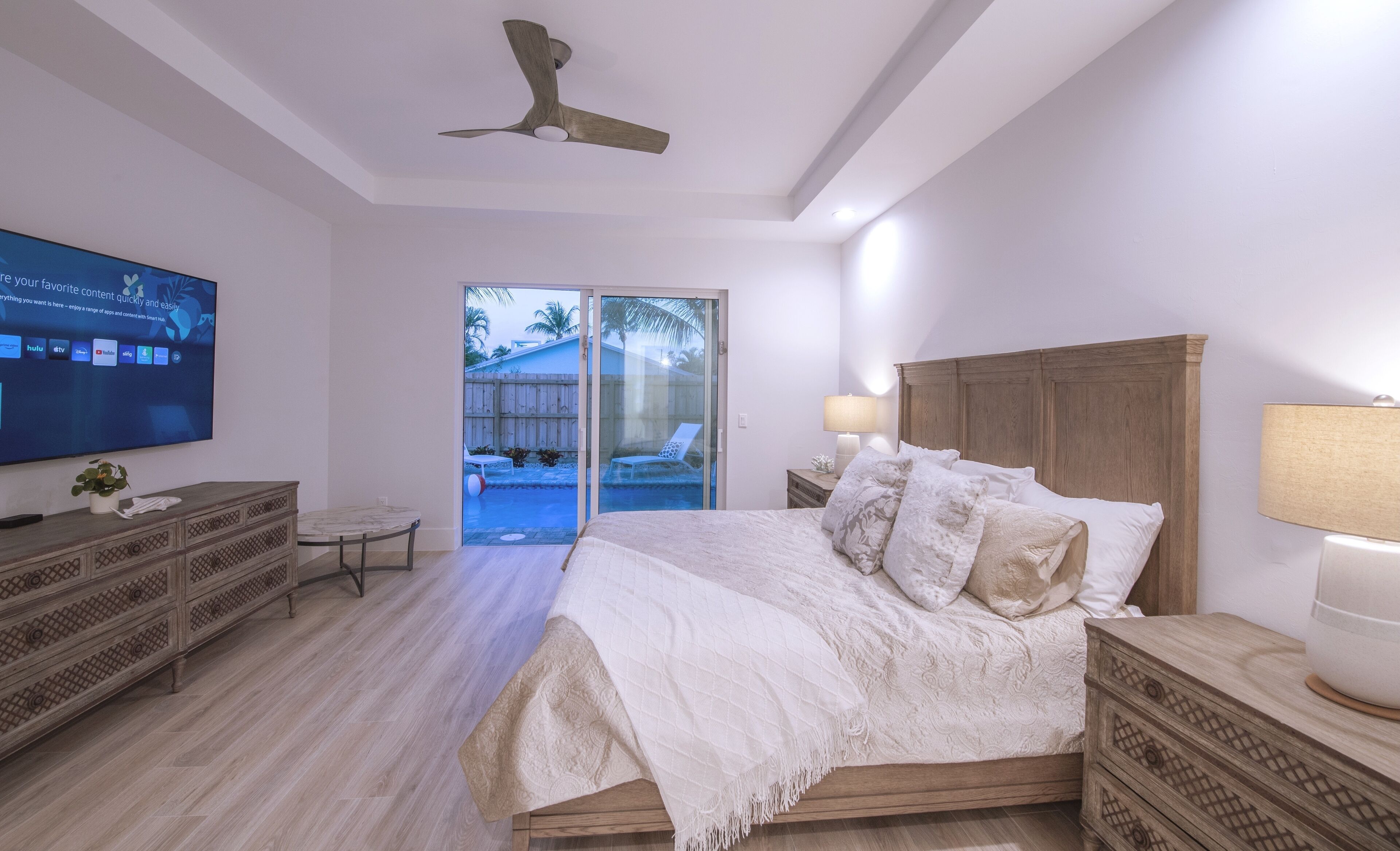 A bedroom with a bed, a TV, a wooden dresser, and a sliding glass door leading to a pool area.