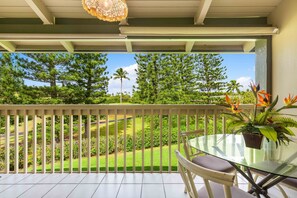 A balcony with a glass table and chairs, overlooking a well-maintained garden with trees and a golf course.
