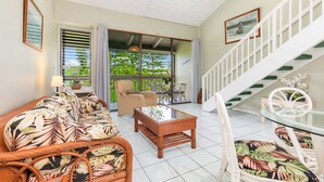 A spacious living area with a glass coffee table, wicker furniture, a staircase with white railings, and a view of greenery outside.