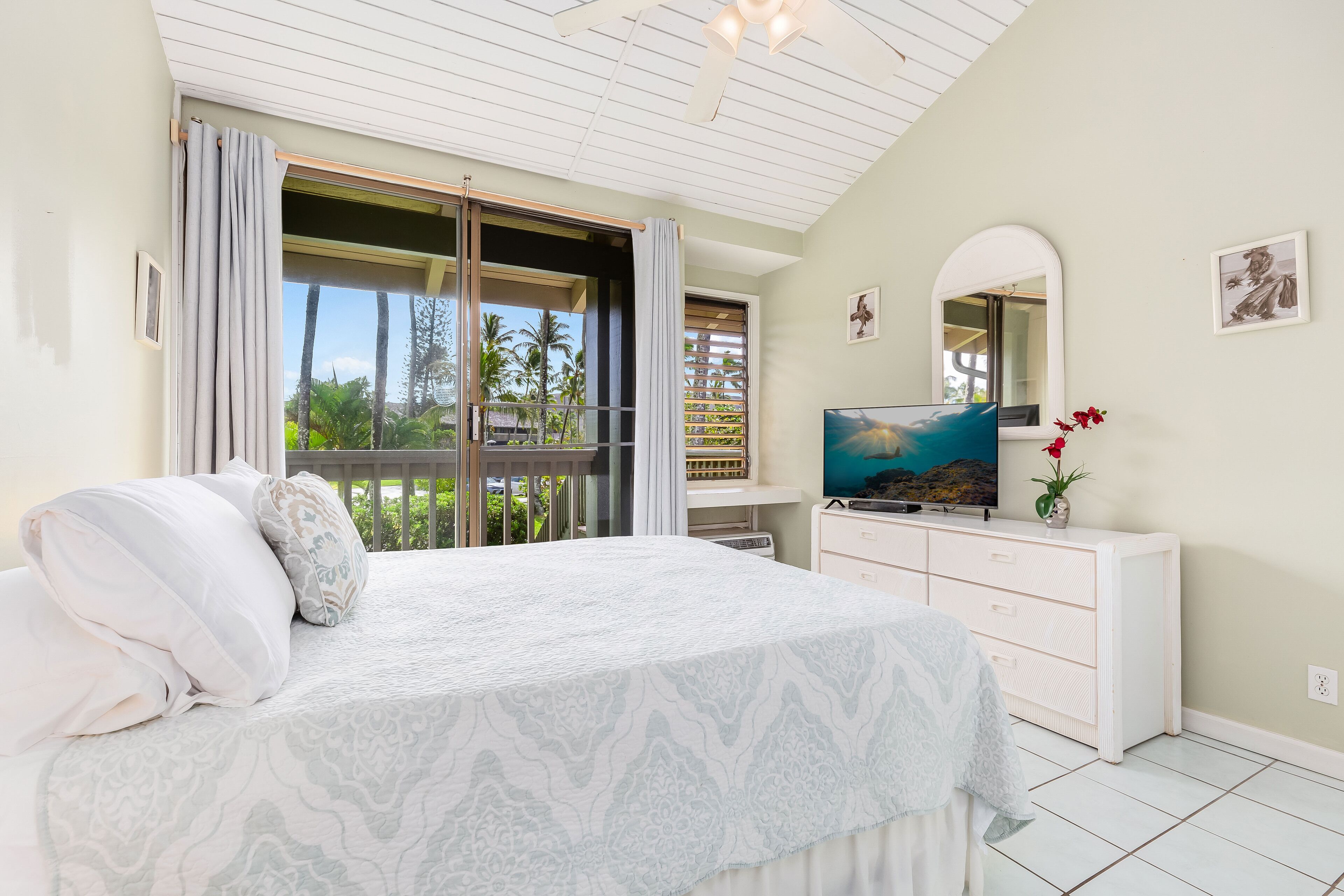 Un dormitorio con cama, televisor, cómoda y balcón con vistas a palmeras.